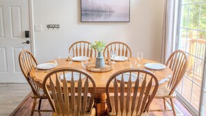 Une salle à manger avec une table en bois dressée pour six personnes, entourée de chaises en bois, un vase avec des plantes et un tableau encadré au mur.