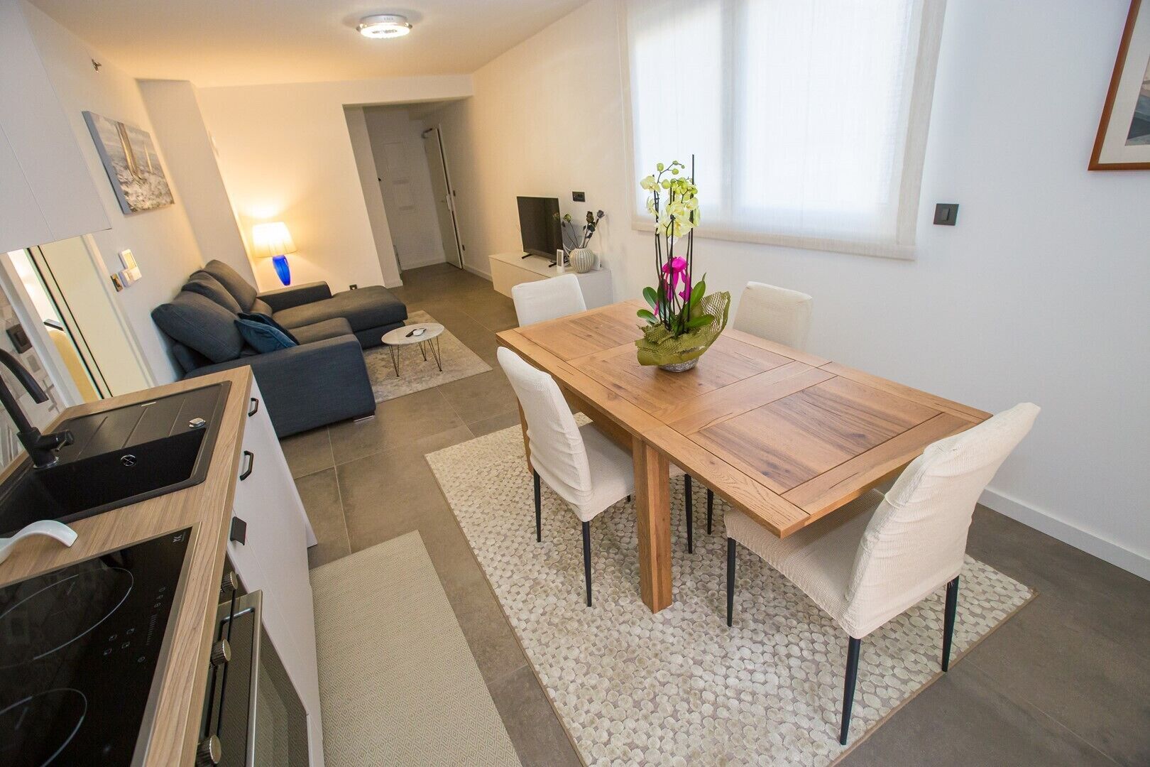 A modern kitchen with a built-in stove and sink, a dining area with a wooden table and white chairs, and a living room with a sofa and a TV.