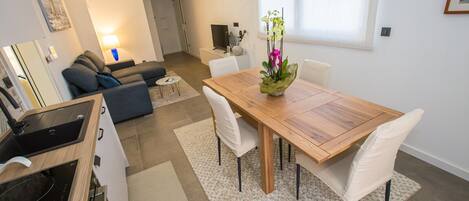 A modern kitchen with a built-in stove and sink, a dining area with a wooden table and white chairs, and a living room with a sofa and a TV.