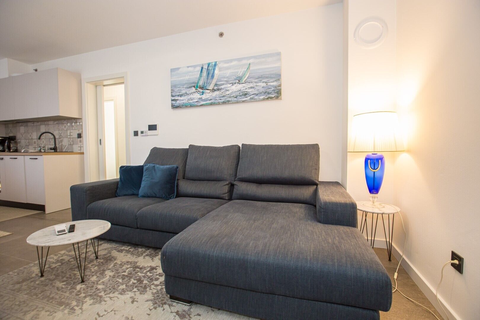 A modern living room with a grey sectional sofa, a small round coffee table, and a blue lamp. A painting of sailboats is on the wall, and a kitchen is visible in the background.
