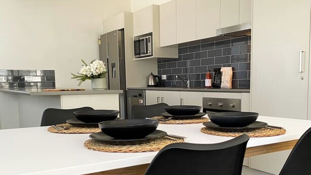 A modern kitchen with white cabinets, a black tiled backsplash, and a white dining table with black bowls and placemats. - 3BR Cottage in the Heart of Bellingen (Bellingen)