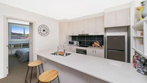 Een moderne keuken met witte kasten, een roestvrijstalen koelkast, een magnetron en een spoelbak. Er staan twee houten krukken en er hangt een klok aan de muur.