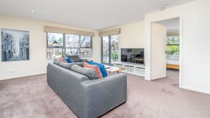 Een moderne woonkamer met een grijze bank, een flatscreen televisie en grote ramen.