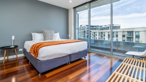 Ein modernes Schlafzimmer mit einem großen Bett, einem kleinen Nachttisch, einem Sessel und einem großen Fenster mit Blick auf die Stadt.