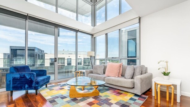 Ein geräumiges Wohnzimmer mit einem großen Glasfenster zum Balkon, einem blauen Samtsofa, einem beigen Sofa, einem Glastisch und einem farbenfrohen Teppich.