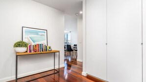 Ein Flur mit Holzboden, einer weißen Wand, einem gerahmten Bild, einem kleinen Tisch mit Büchern und einer Pflanze sowie einer Tür am Ende.