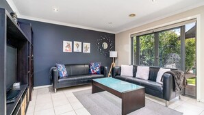 A modern living room with a dark blue wall, two black leather sofas, a glass coffee table, and a large window offering a view of greenery.