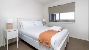 Une chambre à coucher avec un lit, deux tables de chevet, une fenêtre avec un store et une vue sur l’extérieur.