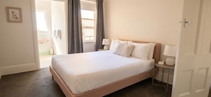 A neatly made bed with white linens, a bedside table with a lamp, and a view of the outside through a window with curtains.