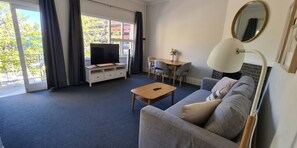 A modern living room with a sofa, a coffee table, a TV on a stand, a dining table with chairs, and a large window with curtains.
