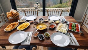 Une table à manger dressée avec différents plats, assiettes et couverts, offrant une vue panoramique à travers de grandes fenêtres.