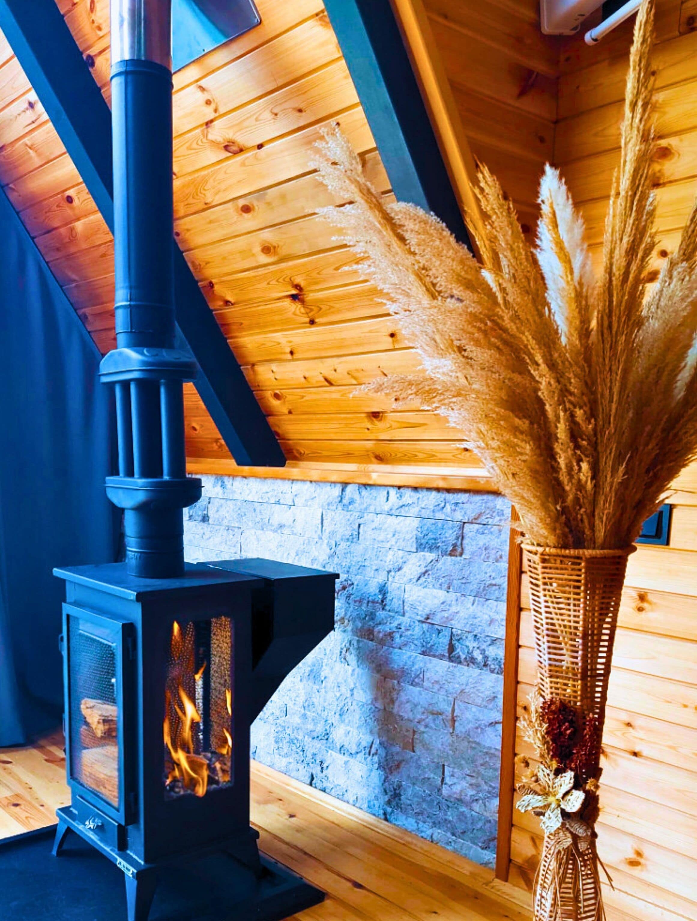 A wood-burning stove with a chimney, a stone wall, and a vase with pampas grass.