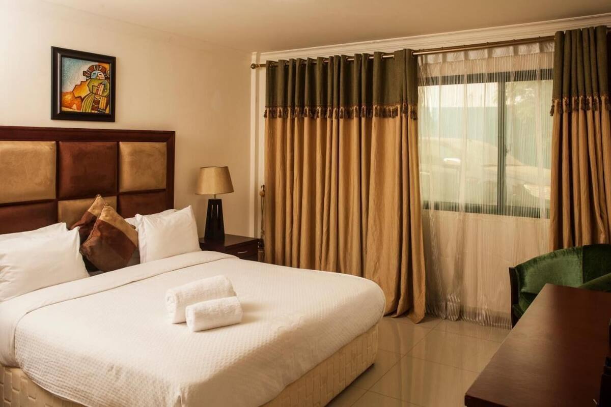 Une chambre d’hôtel avec un grand lit, une tête de lit, un bureau, une chaise, une lampe, des rideaux et un tableau au mur.