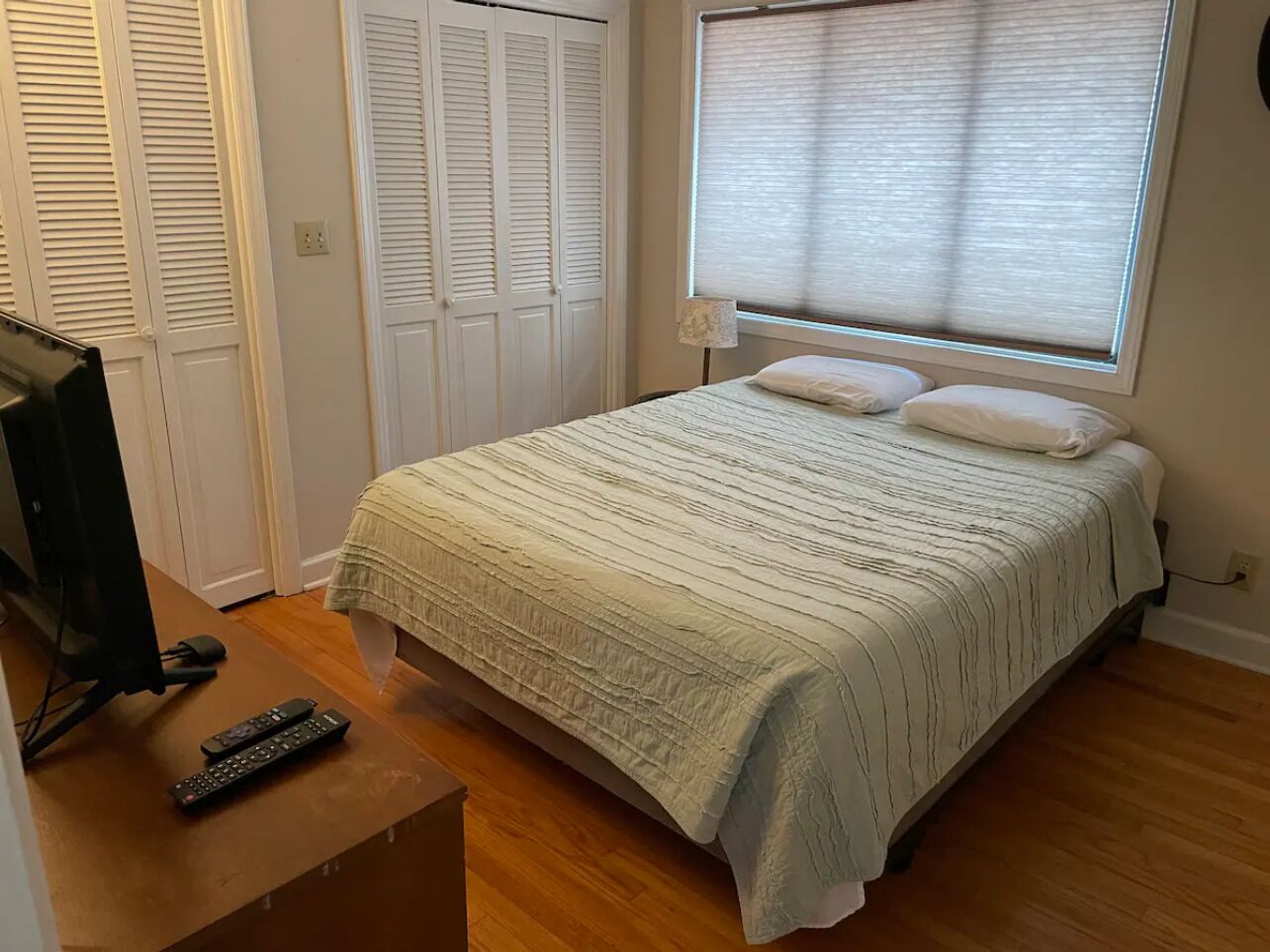 Une chambre à coucher avec un lit, un téléviseur posé sur une table en bois et une fenêtre avec des stores.