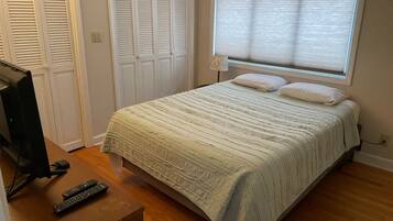 Une chambre à coucher avec un lit, un téléviseur posé sur une table en bois et une fenêtre avec des stores.