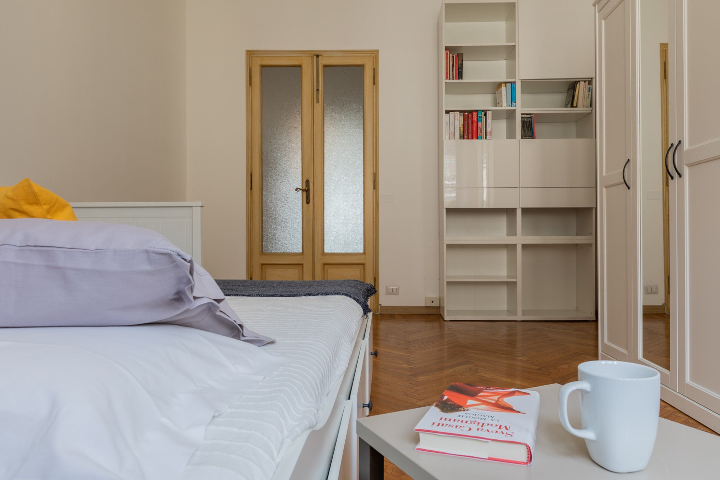 A bedroom with a bed, a bedside table with a book and a mug, a wardrobe, and a door with frosted glass panels.