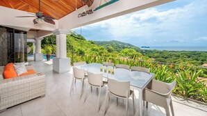 A modern outdoor patio with a dining table and chairs, a wicker sofa with orange cushions, and a ceiling fan. - Casa del Mar • 4 BR Ocean-View Villa with Private Pool & BBQ • 8 Guests (Playa Hermosa)