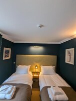 Habitación de hotel con dos camas, cabecera, mesita de noche con lámpara y obras de arte en las paredes.