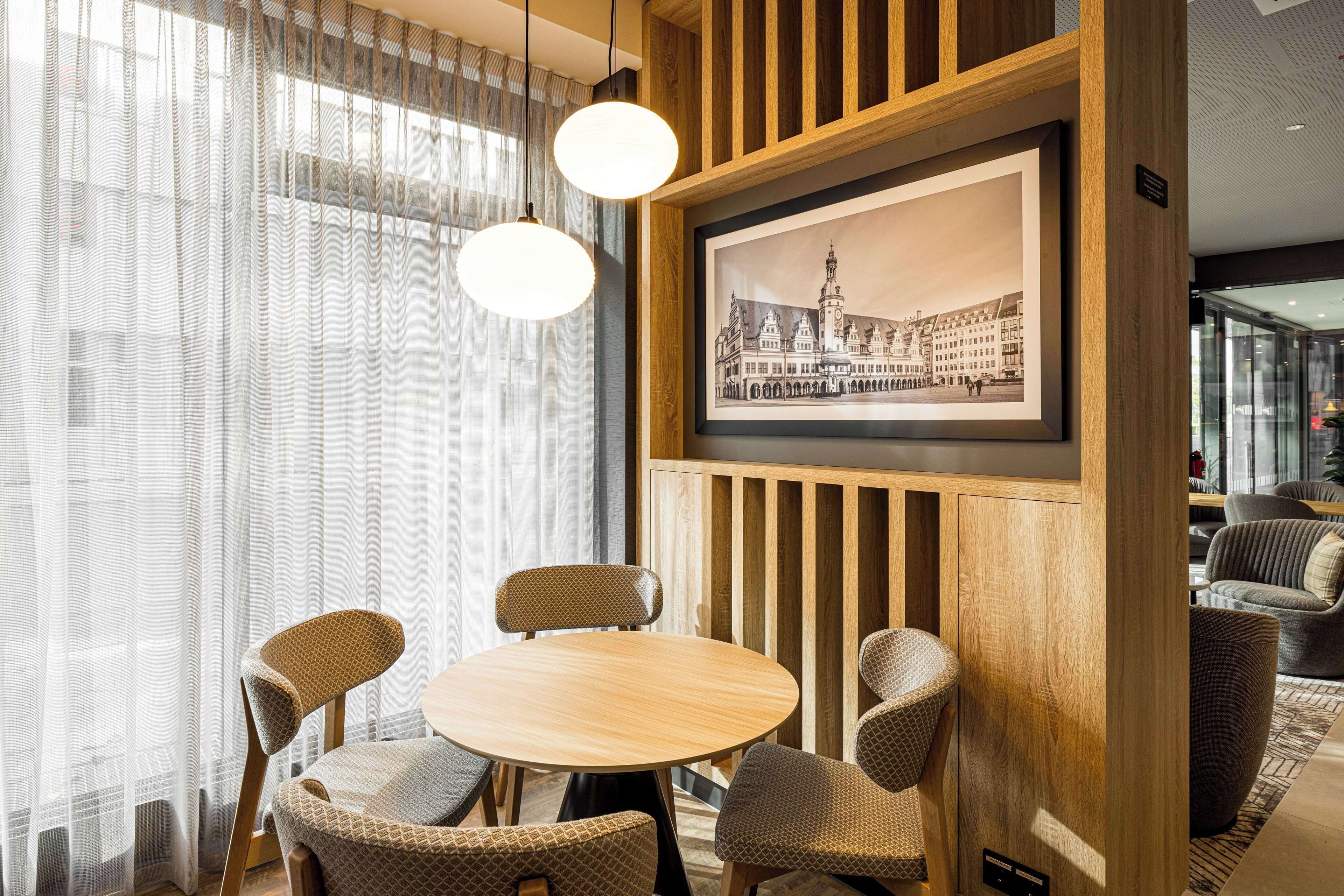 A dining area with a round wooden table and four patterned chairs, a framed black and white cityscape picture on the wall, and sheer curtains on the windows.