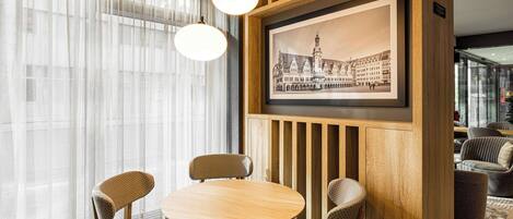 A dining area with a round wooden table and four patterned chairs, a framed black and white cityscape picture on the wall, and sheer curtains on the windows.
