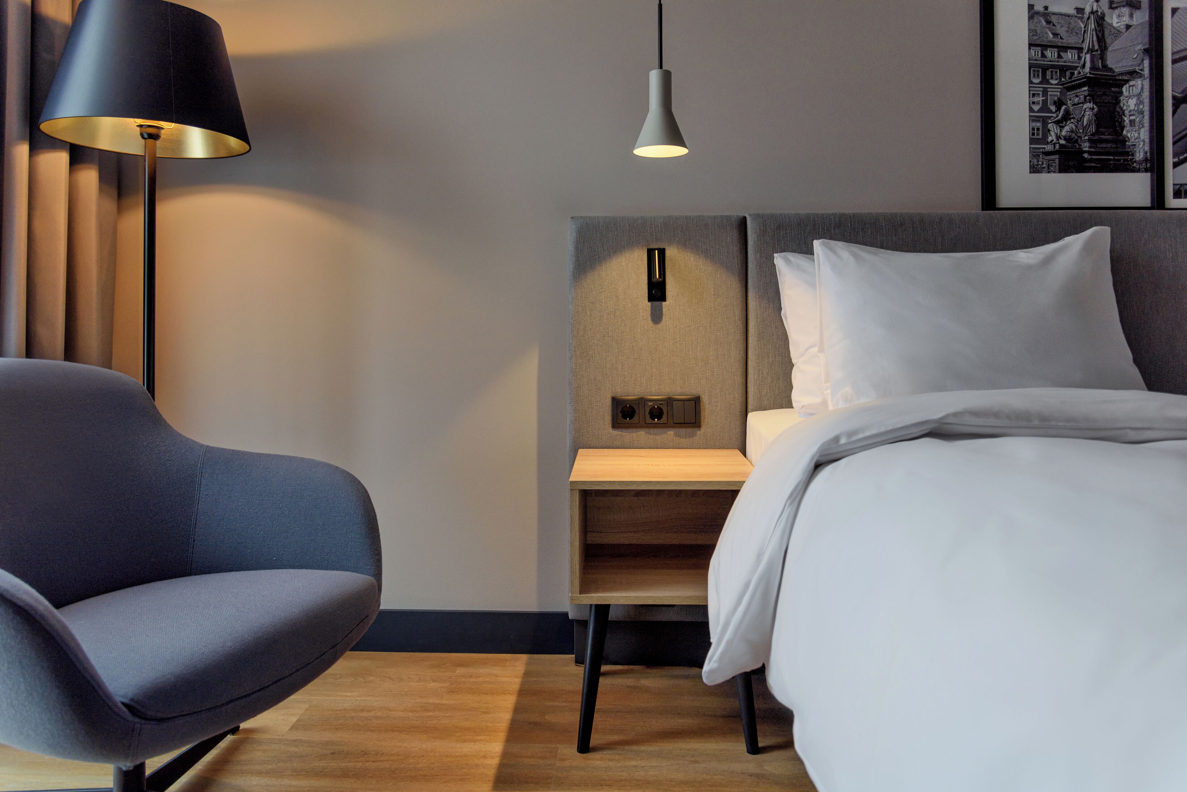 Una habitación de hotel moderna con una cama, una silla, una mesita de noche, una lámpara y una lámpara de pared.
