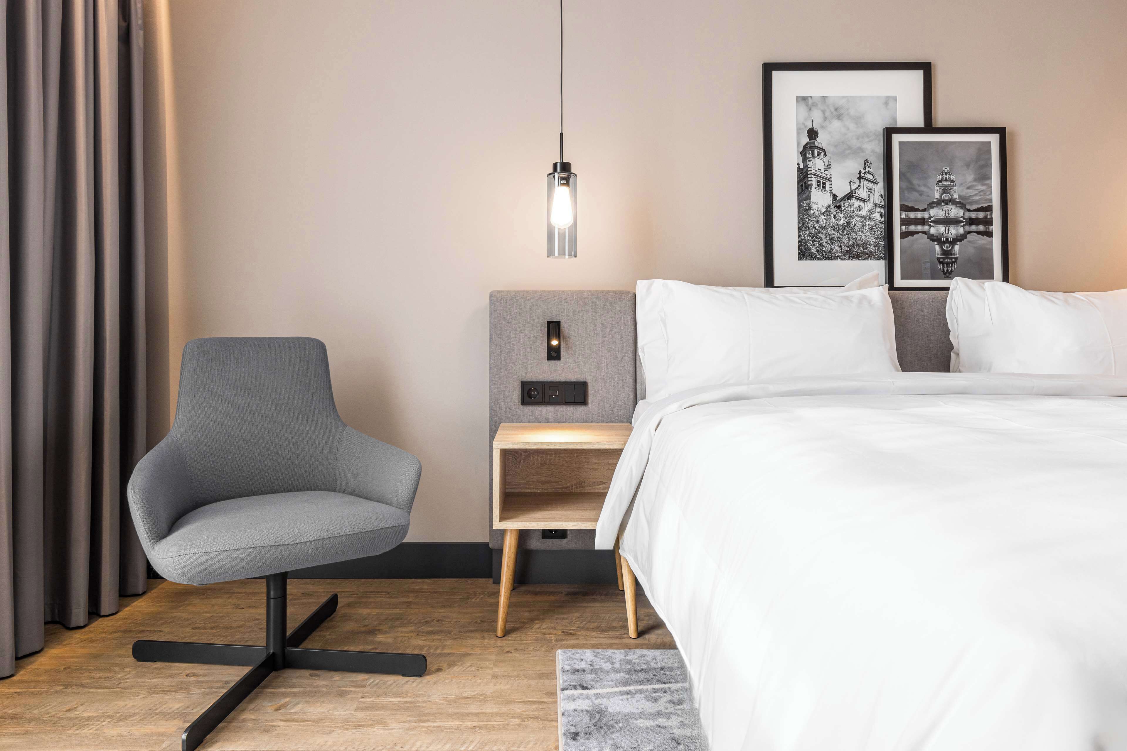 A modern hotel room with a grey armchair, a bedside table with a lamp, a bed with white linens, and two framed pictures on the wall.