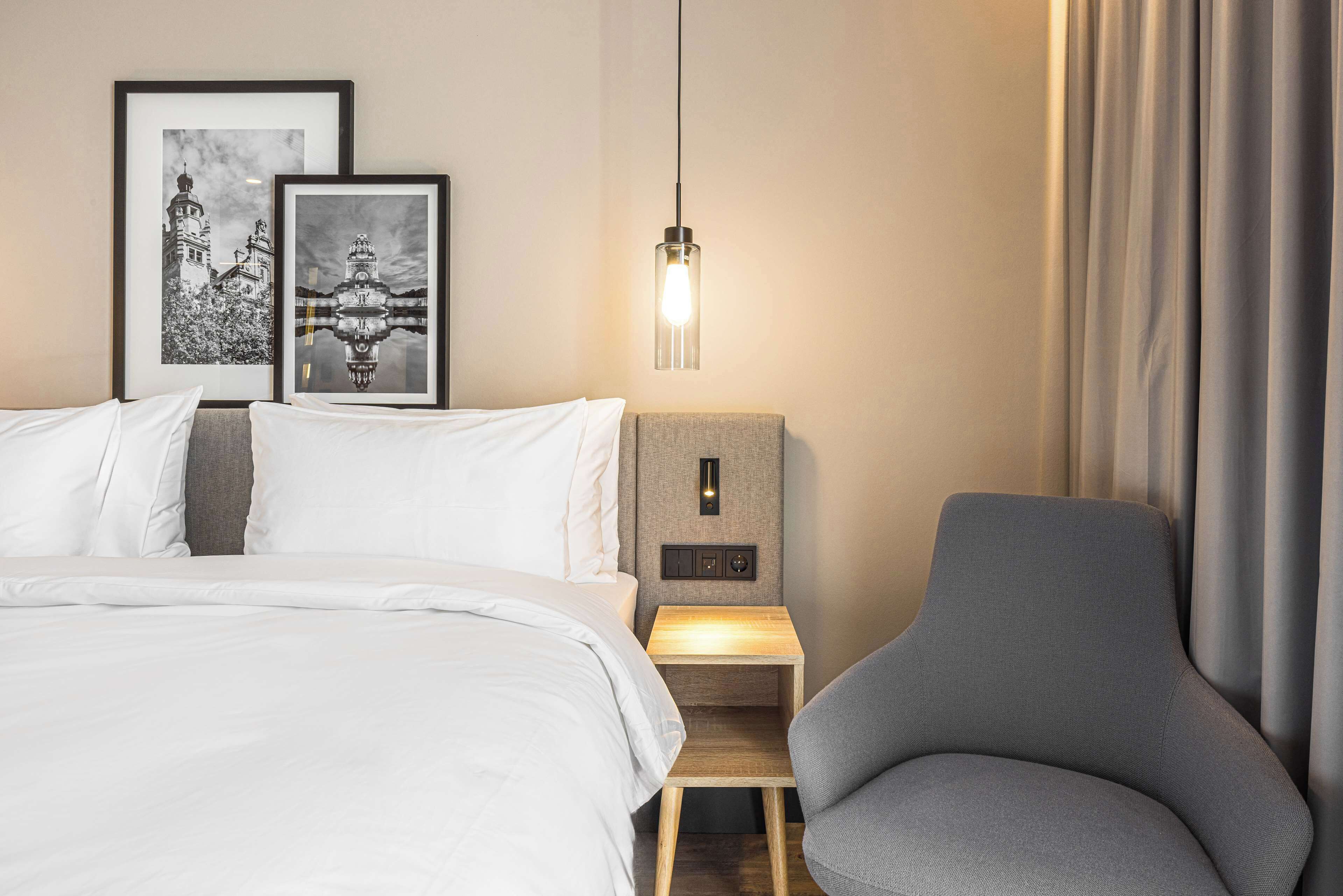 Et pænt og ryddeligt hotelværelse med seng, stol, natbord med lampe og to billeder i rammer på væggen.