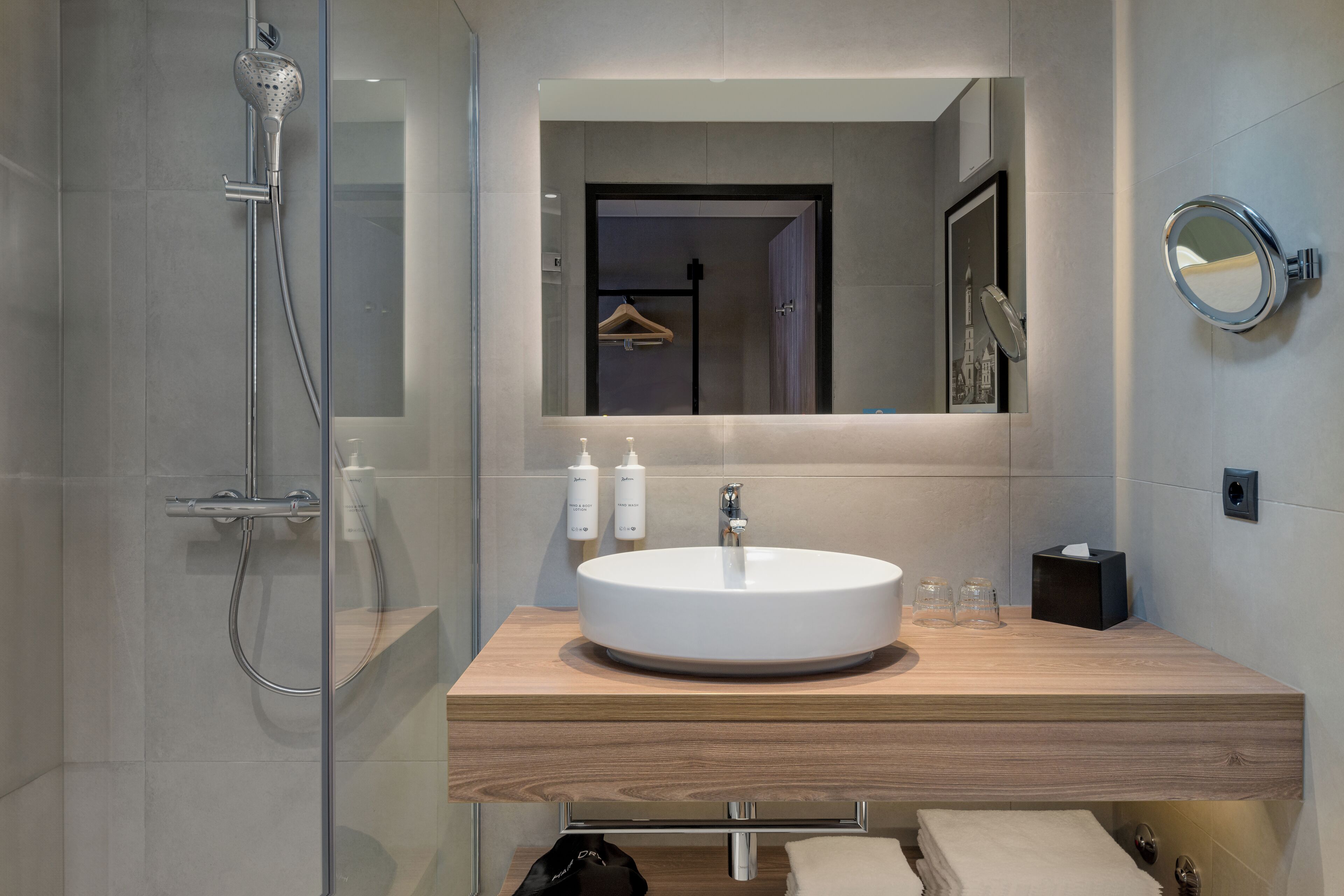 Un baño moderno con cabina de ducha de vidrio, tocador de madera, espejo redondo y lavabo.