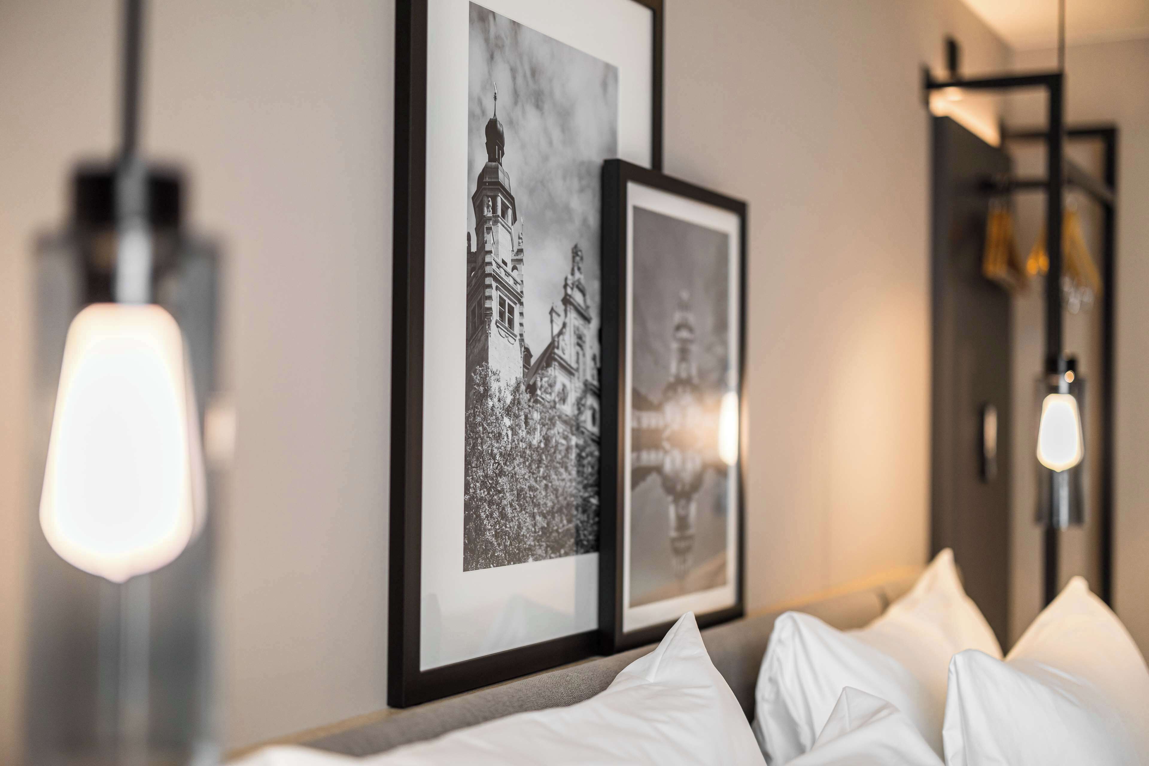 A hotel room with a bed, white pillows, and two framed black and white photographs on the wall.