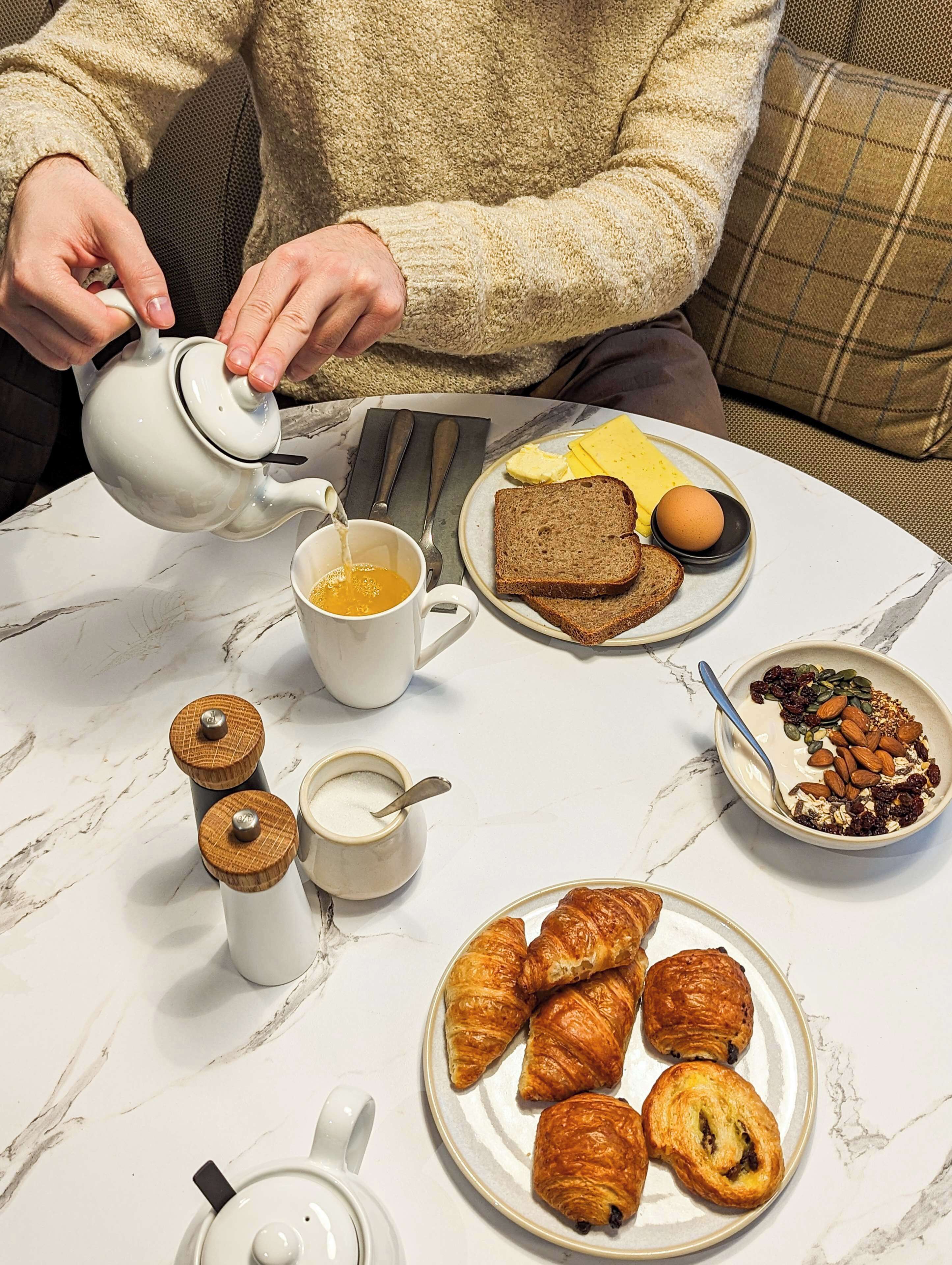 A breakfast setting with a teapot pouring tea, bread, eggs, and pastries on a marble table.
