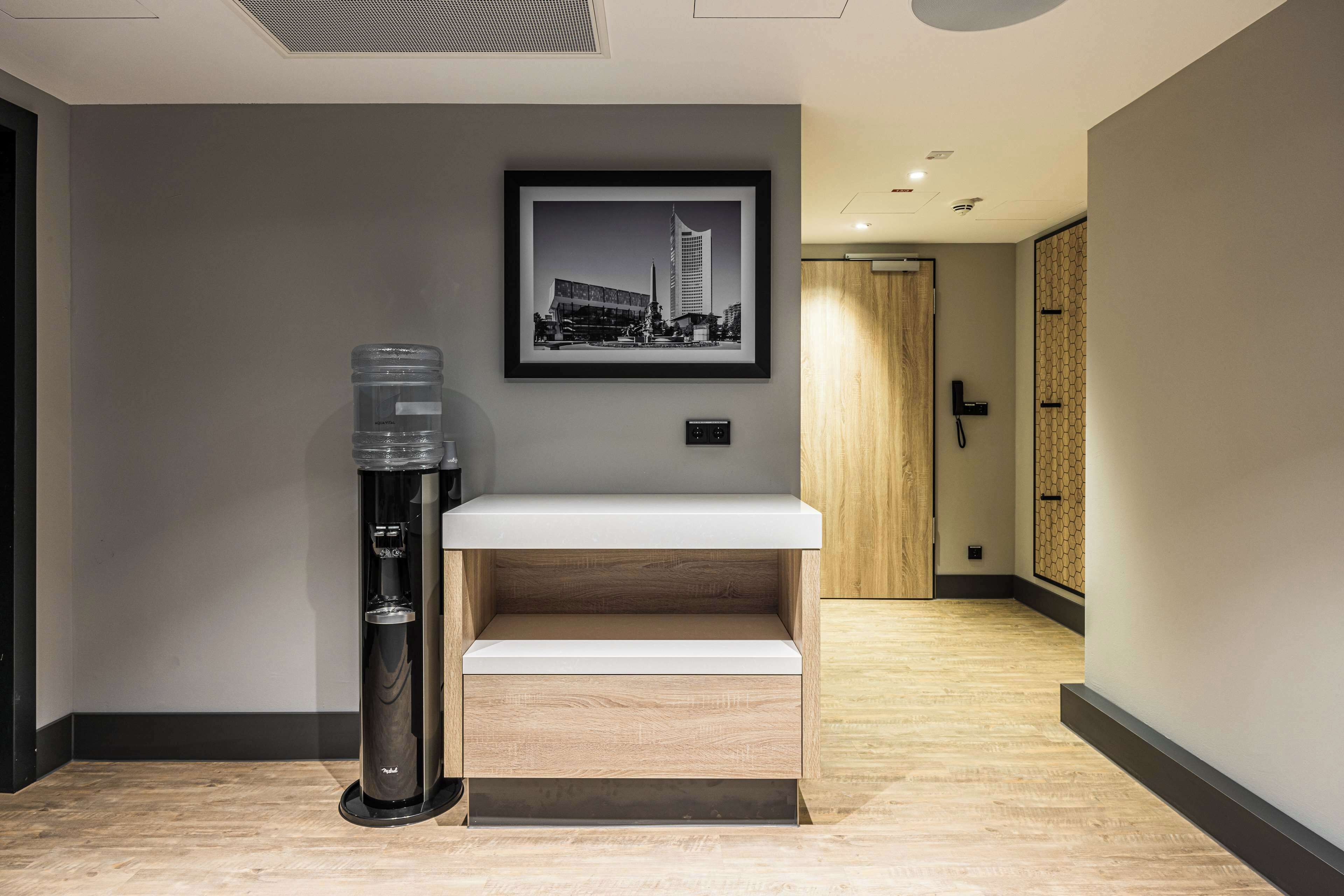 Zona de recepción de un hotel con dispensador de agua, una fotografía enmarco de un paisaje urbano, mostrador de recepción y una puerta de madera al fondo.
