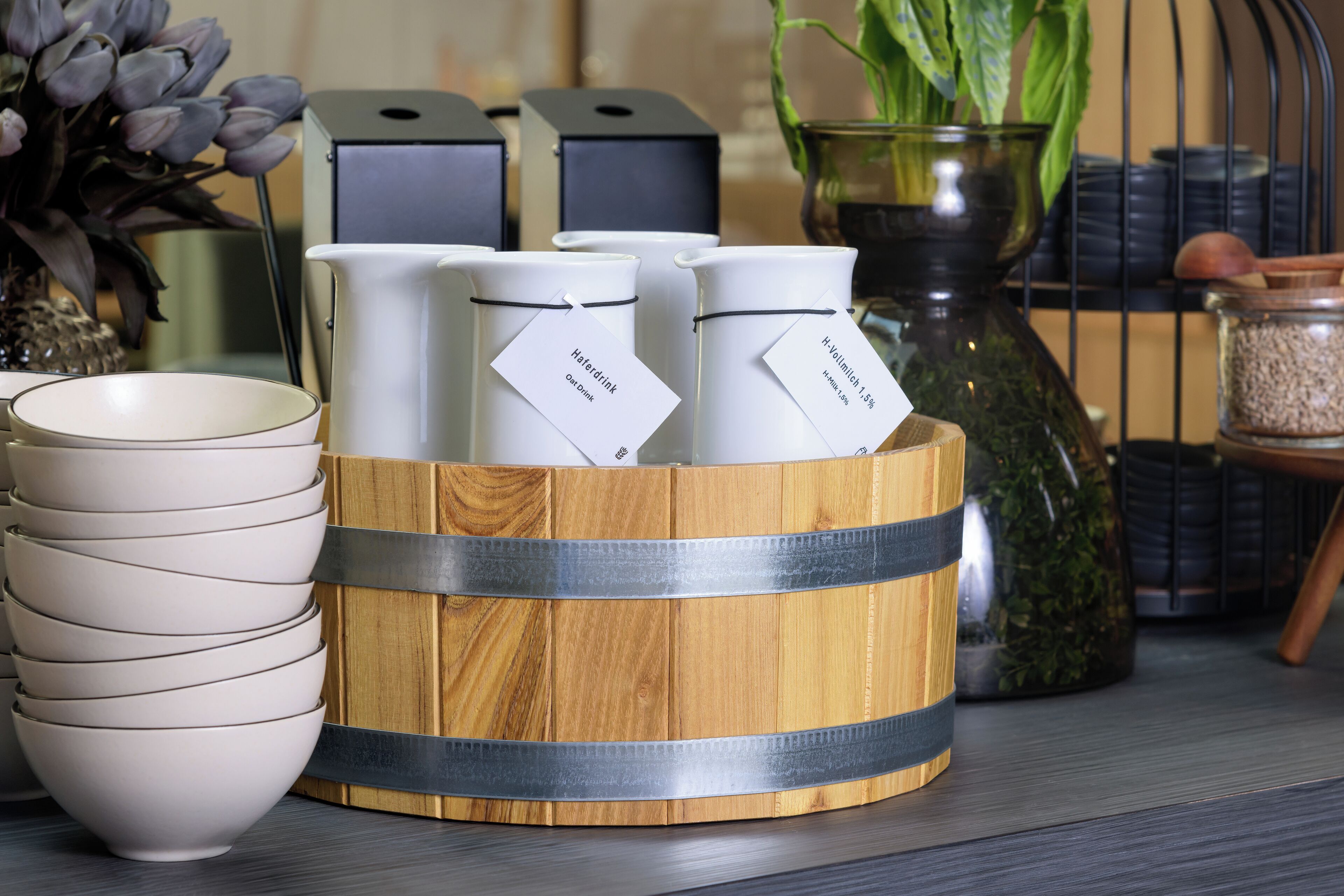 A wooden basket holding white containers with tags, a stack of white bowls, and a vase with green leaves.