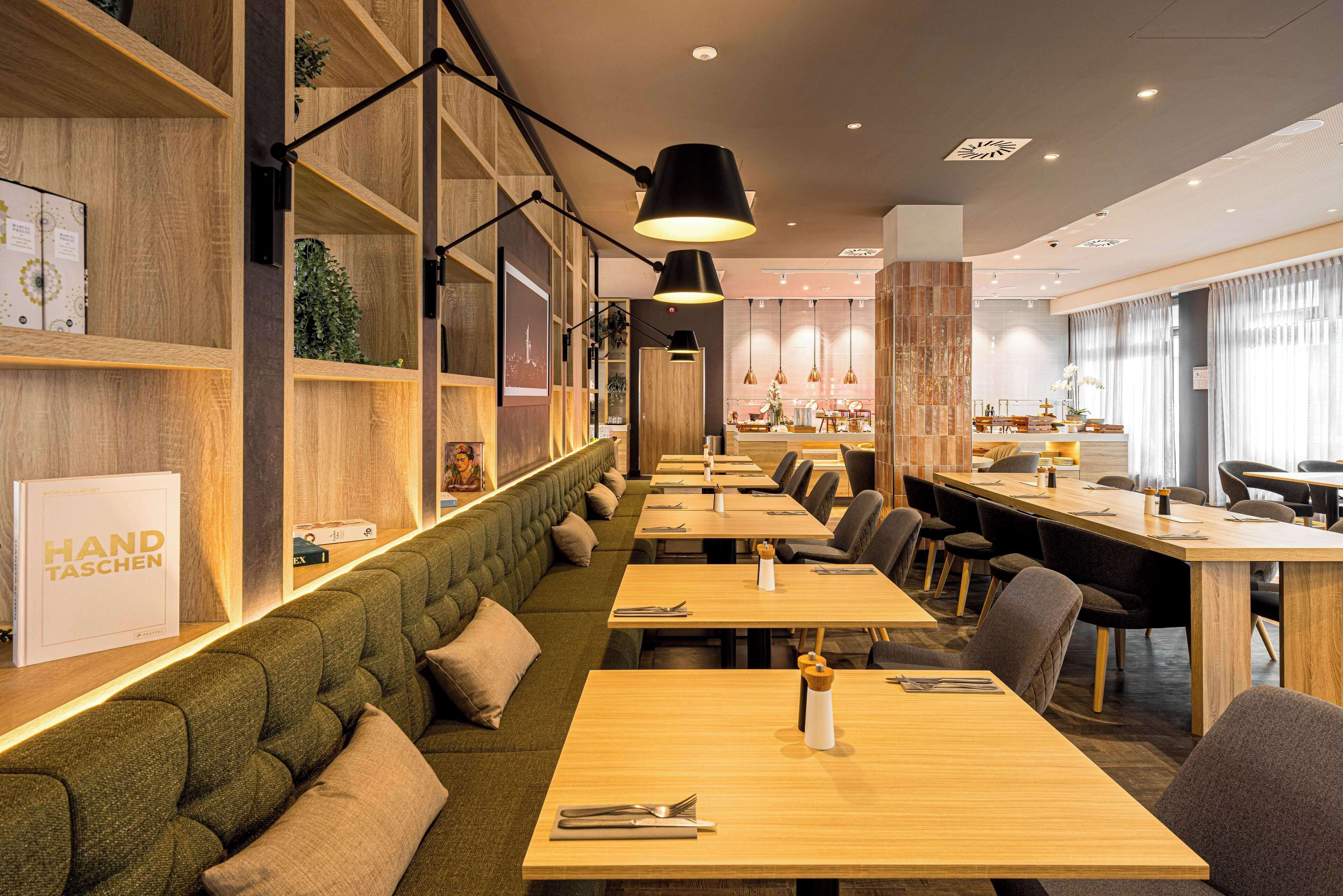 A restaurant with long wooden tables, green cushioned booths, and pendant lighting.