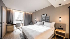 Habitació d'hotel moderna amb un llit gran, una taula de treball amb una cadira, una taula de nit i una finestra gran amb cortines.
