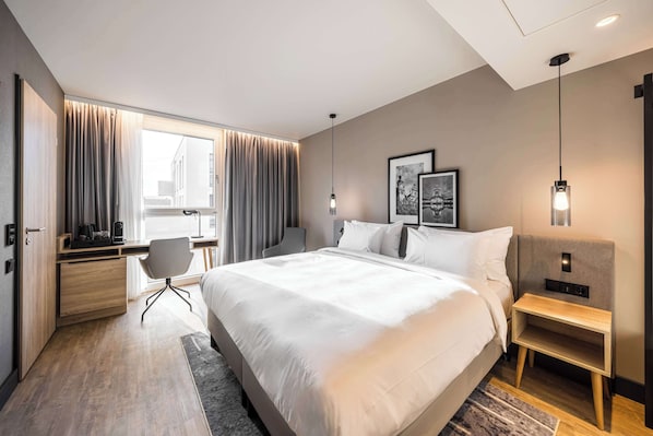 A modern hotel room with a large bed, a desk with a chair, a bedside table, and a large window with curtains.