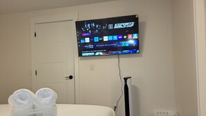 Habitación de hotel con cama, televisor en la pared mostrando la Esports World Cup, aspiradora y cesto de ropa.