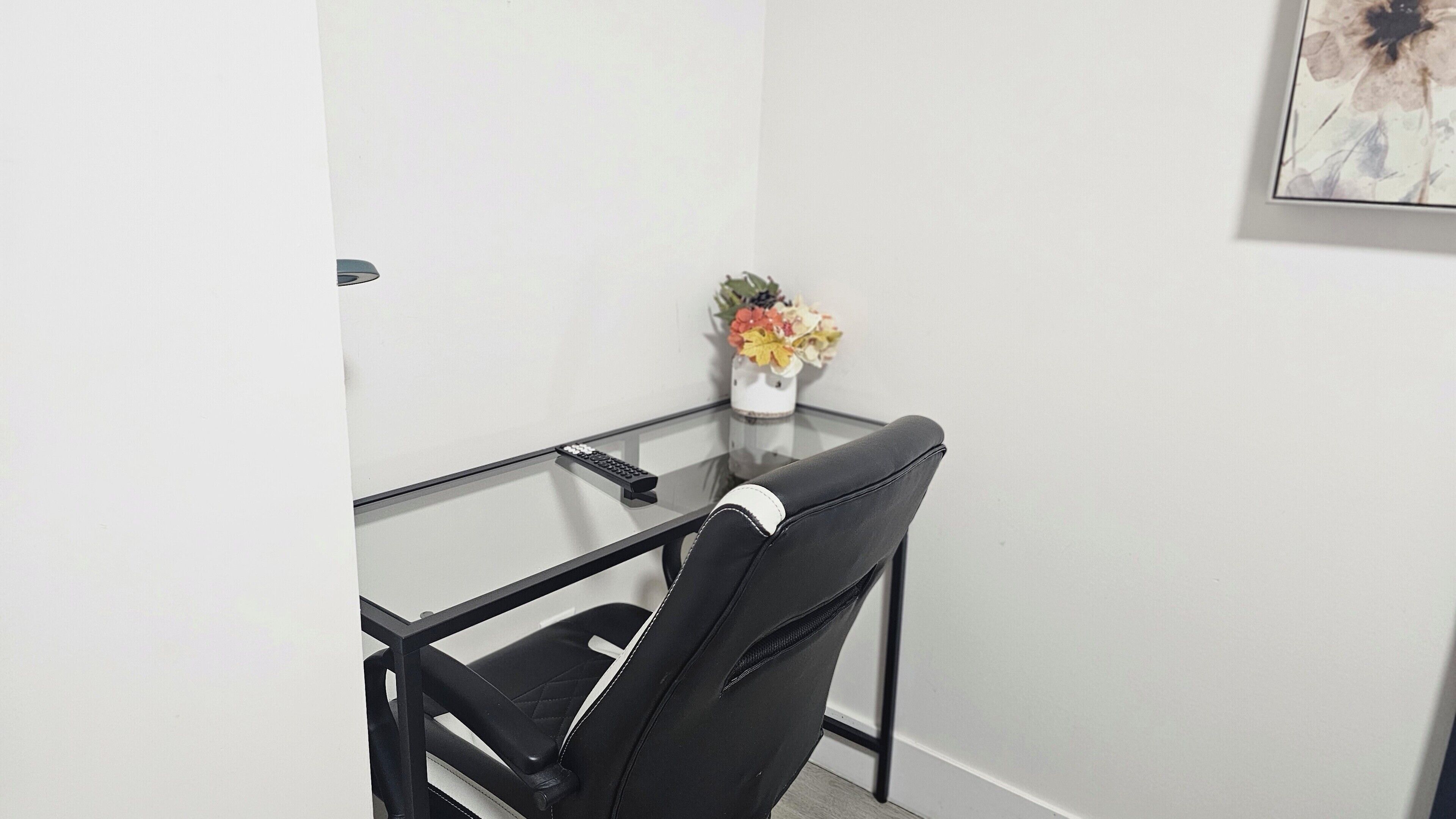 A small office space with a glass table, a black office chair, and a vase with flowers on a white wall.