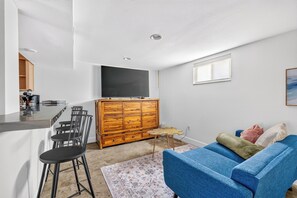 A compact living space with a kitchenette, a wooden cabinet, a flat-screen TV, a blue sofa, and a patterned rug.