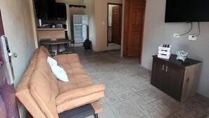 Una sala de estar con un sofá, un televisor montado en la pared, un ventilador de techo y una cocina al fondo.