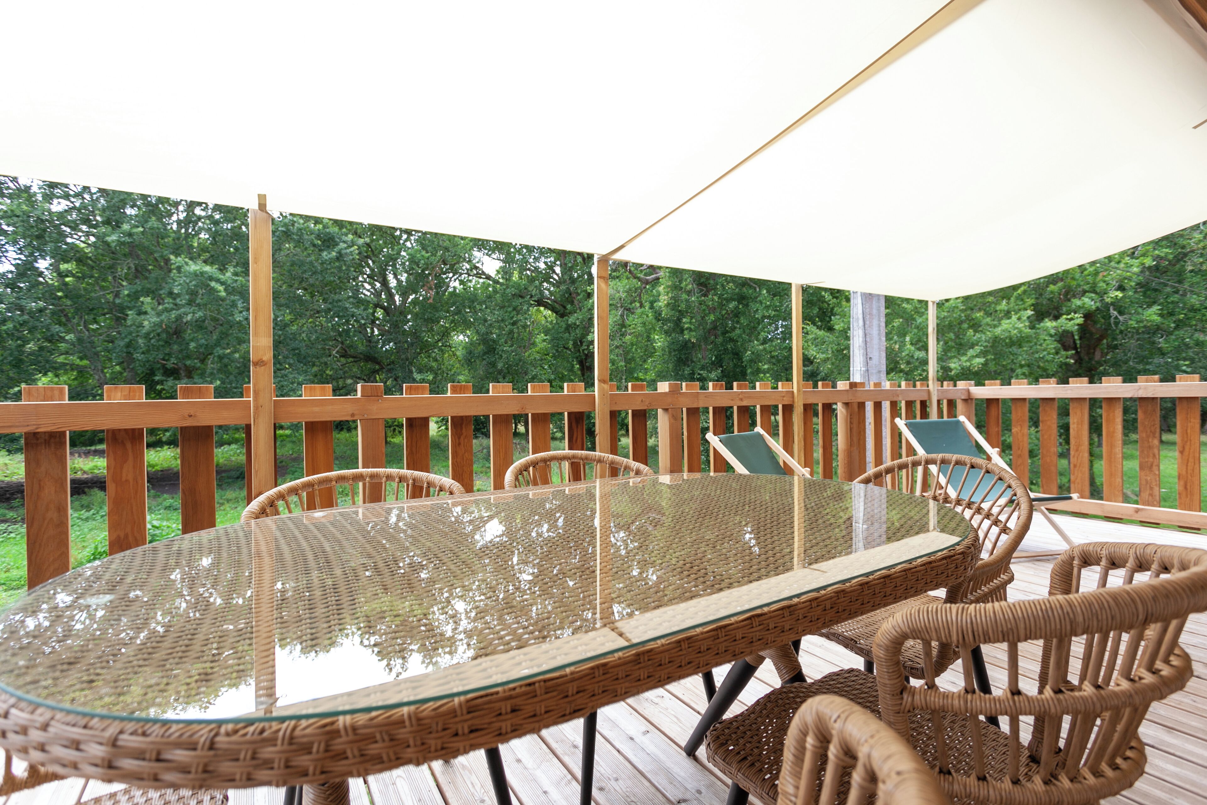 An outdoor deck with a glass-top table and wicker chairs, surrounded by a wooden fence and greenery.