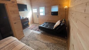 A cozy cabin room with a fireplace, a black leather sofa, a television mounted on the wall, and a small window. - The Cabin at the lodge (Sodus Point)