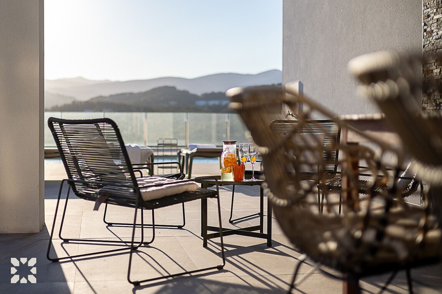 A balcony with a view of mountains, a table with drinks, and two chairs.