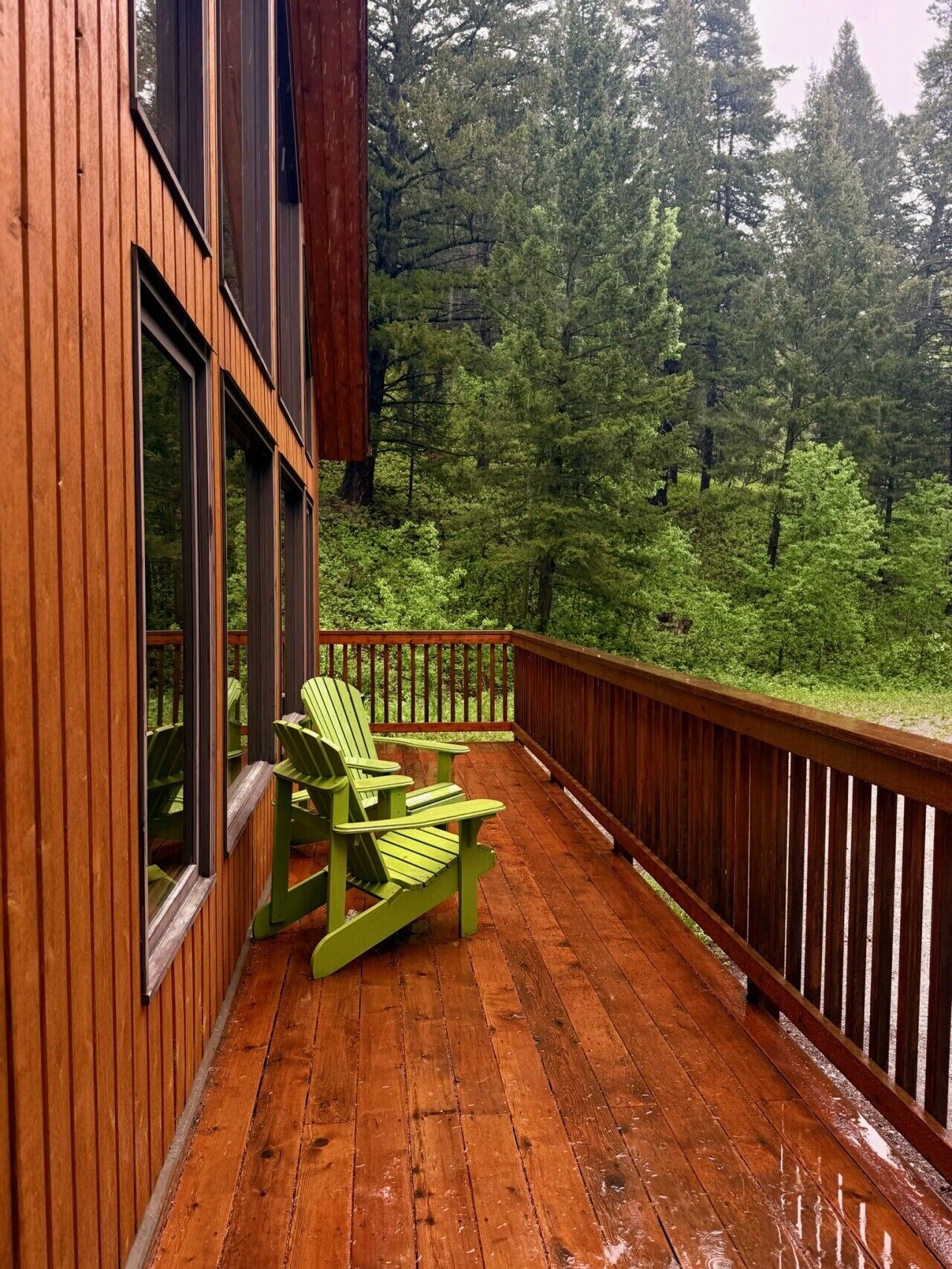 Een houten terras met een groene Adirondack-stoel, omringd door bebossing.