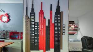 A room with a bed, a chair, a wall clock, and a wall-mounted artwork of skyscrapers.