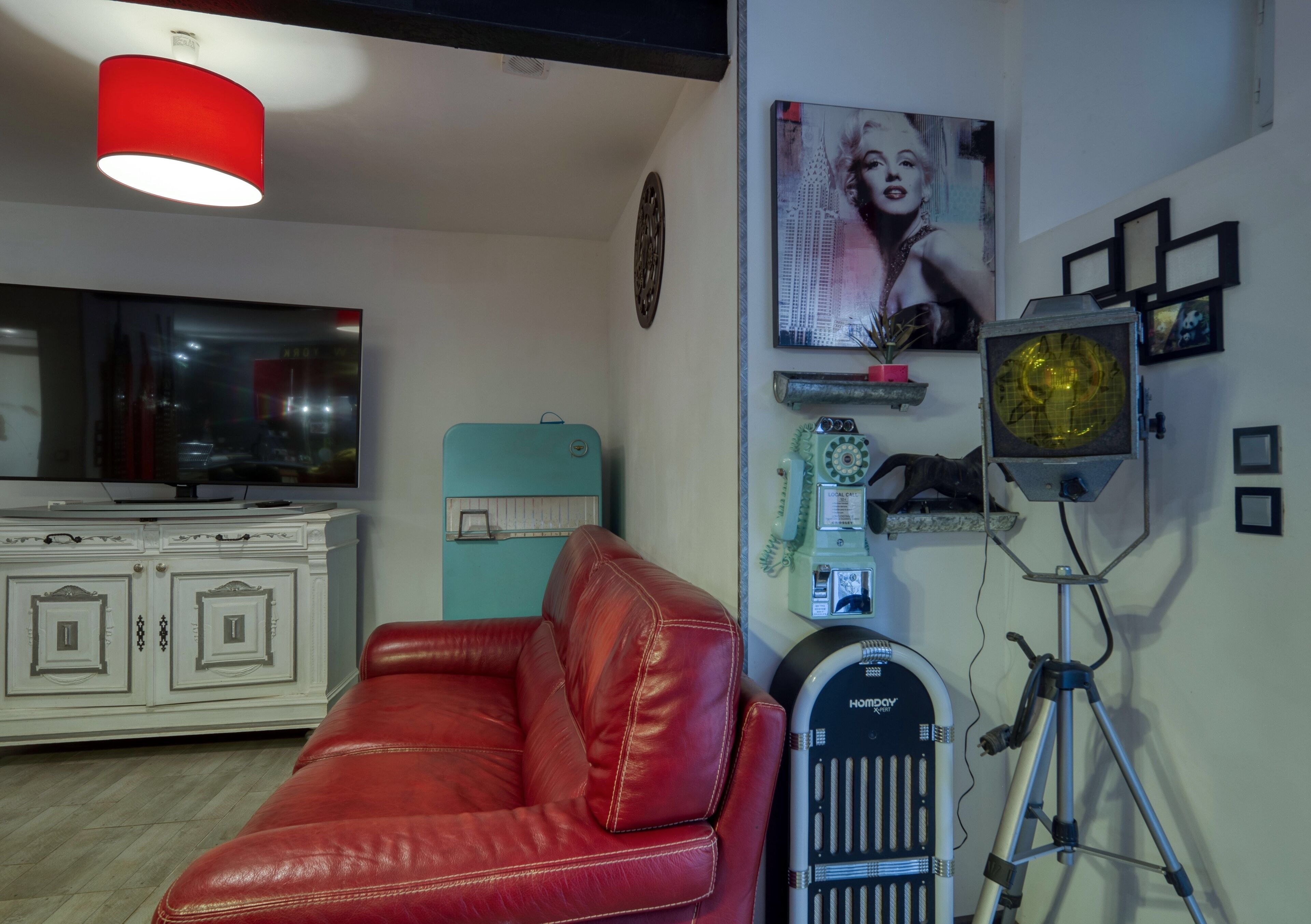 A room with a red leather sofa, a white cabinet, a red pendant light, a wall-mounted TV, a vintage radio, and a tripod with a light.