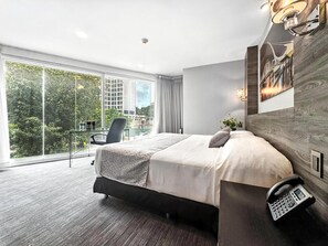 A modern hotel room with a large bed, a desk with a chair, a phone, and a view of the city through large windows. - Elite 47 Unity (Huixquilucan)