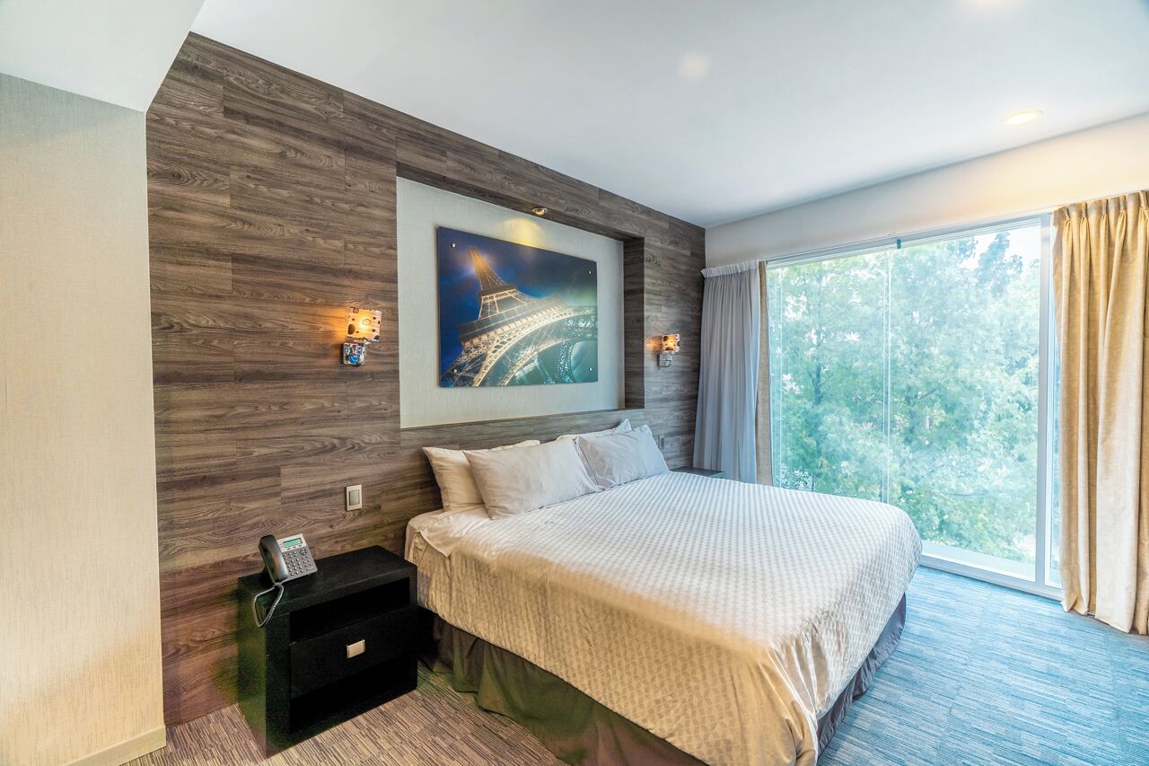 A modern hotel room with a large bed, a painting of the Eiffel Tower, a nightstand with a phone, and a window with curtains.