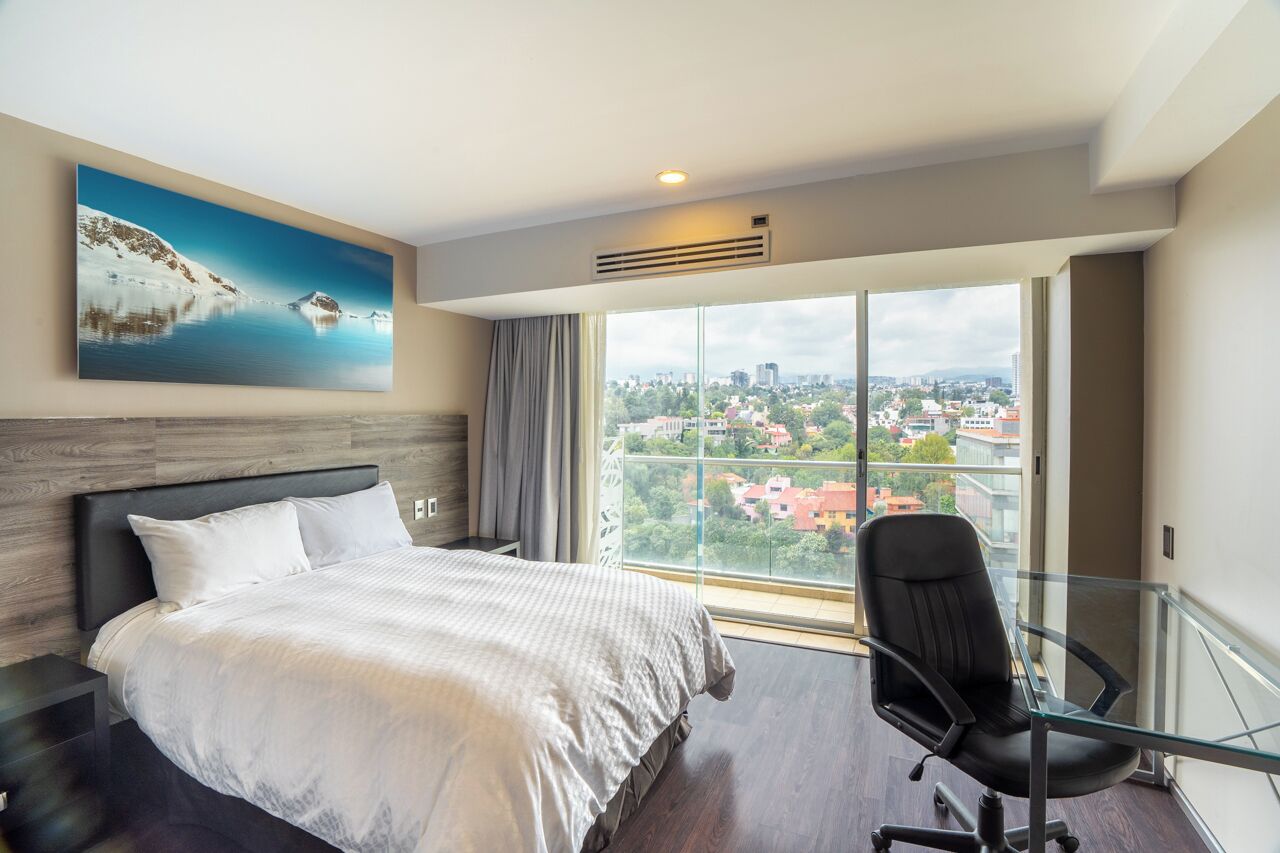 A hotel room with a large bed, a glass desk, a city view, and a painting on the wall.