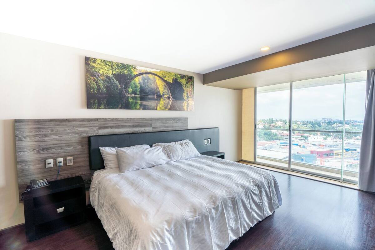 Quarto de hotel moderno com uma cama grande, uma mesinha de cabeceira, telefone fixado na parede e uma janela ampla com vista para a cidade.