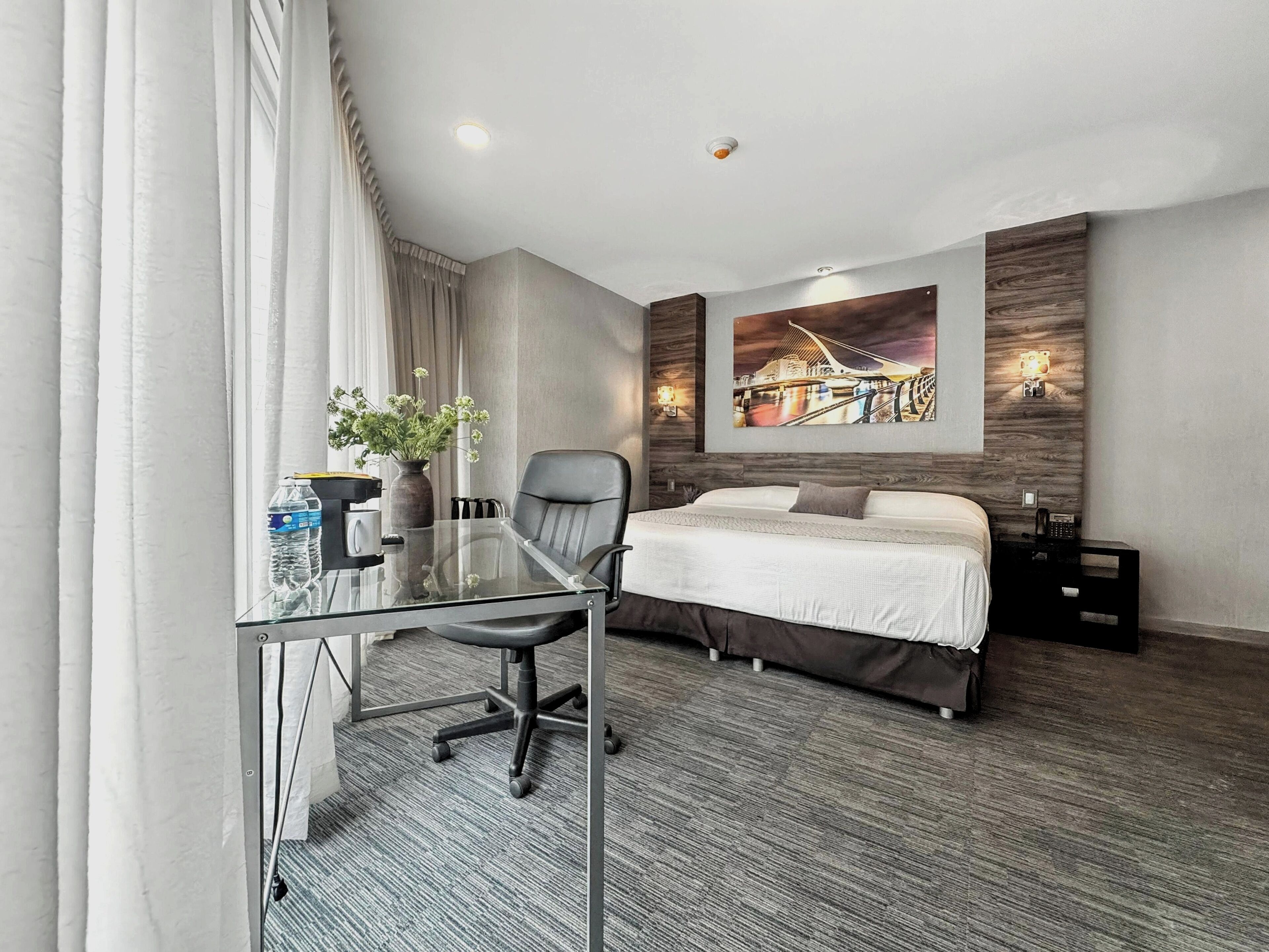 Habitación de hotel con escritorio de vidrio, cama, mesita de noche y un cuadro en la pared.